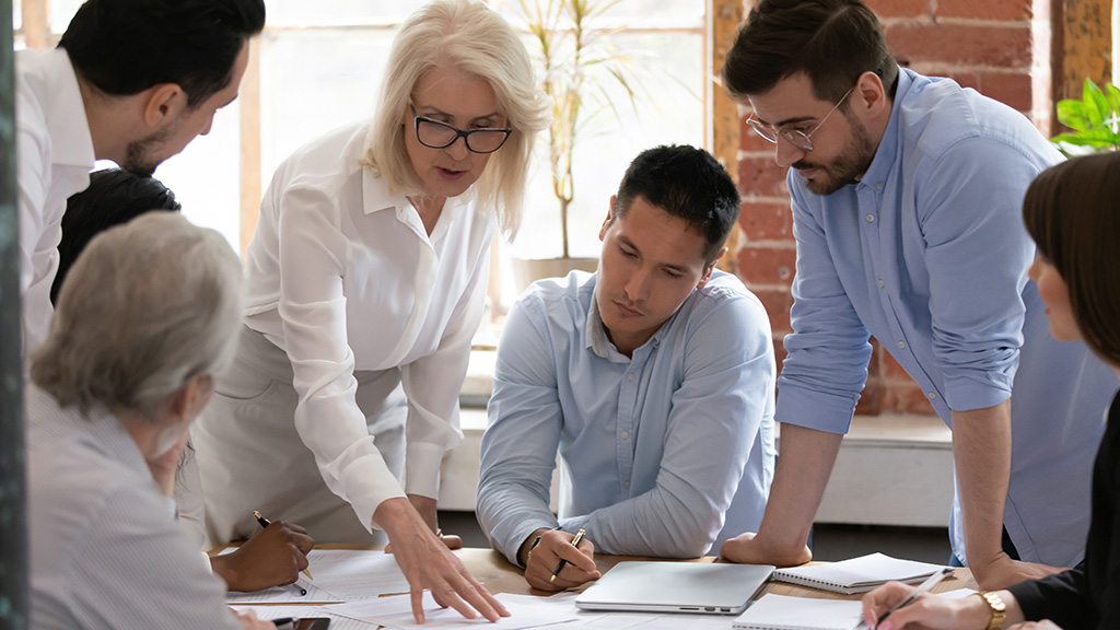 Serious old mature woman team leader coach teach young workers explain paper business plan at group meeting, focused senior female teacher mentor training diverse staff at corporate office workshop